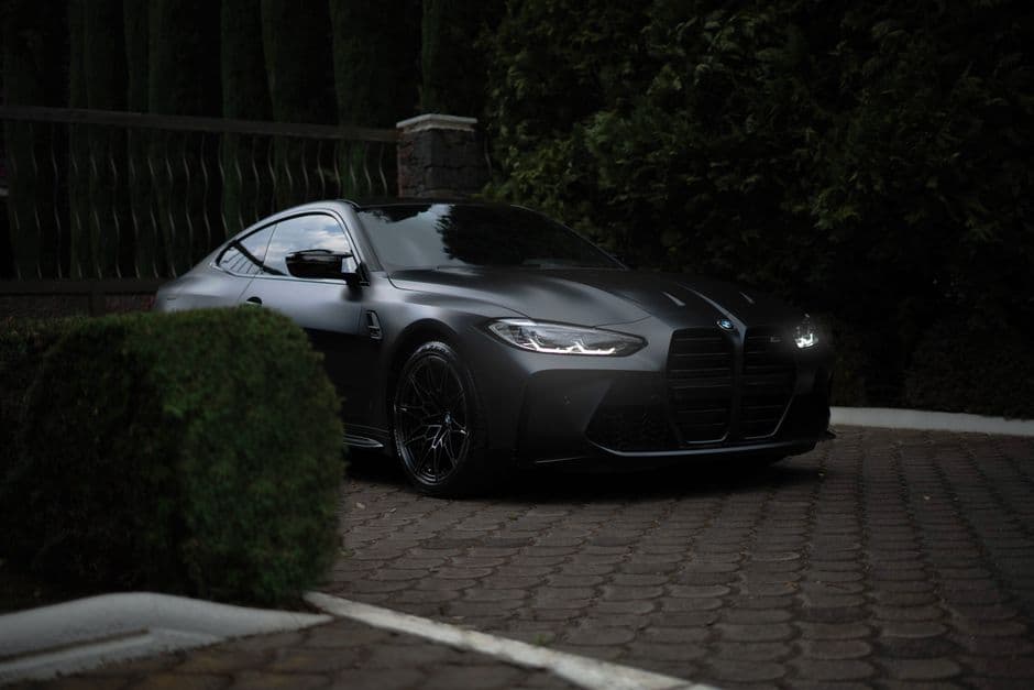 Mat donkergrijze BMW-coupé op klinkers tussen hagen en bomen