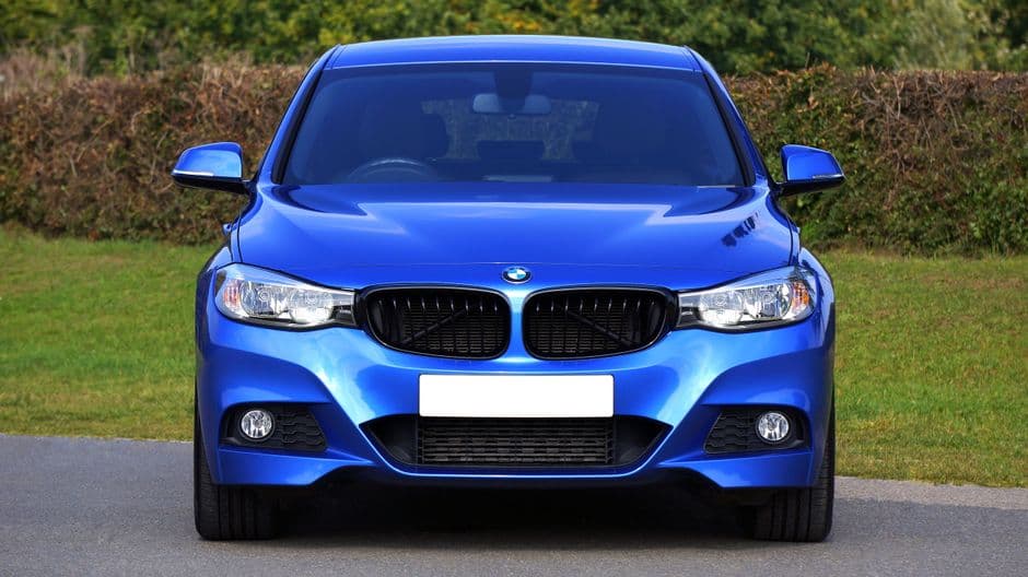 Blauwe BMW recht van voren op een weg met een haag erachter